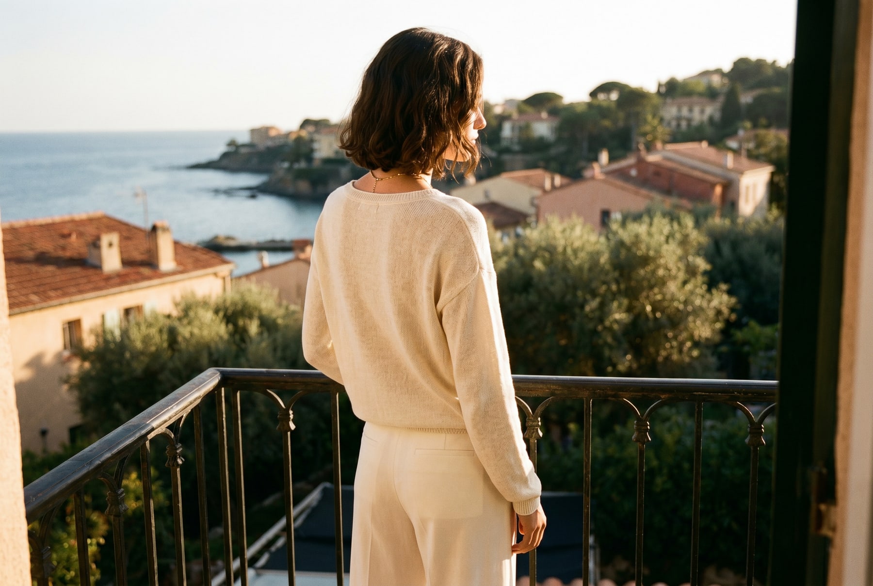 A woman on a Mediterranean balcony at golden hour, leaning on a wrought-iron rail, looking out over the coast.