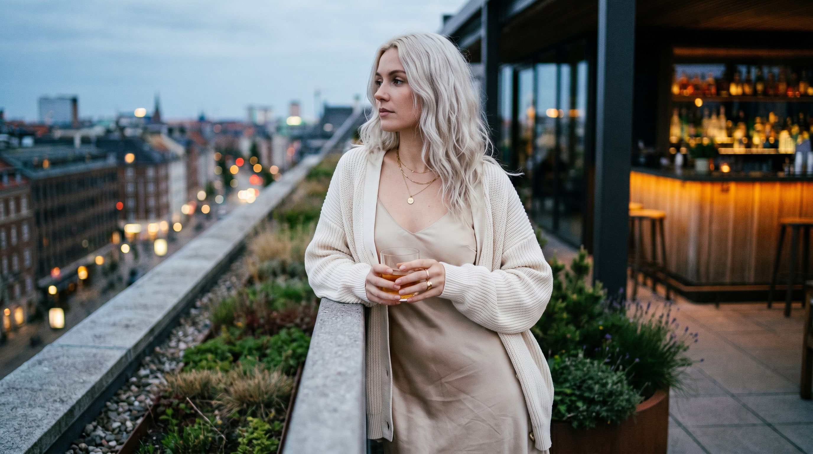 A creator on a rooftop at dusk with a cocktail, evening dress, city blurred behind in blue hour.