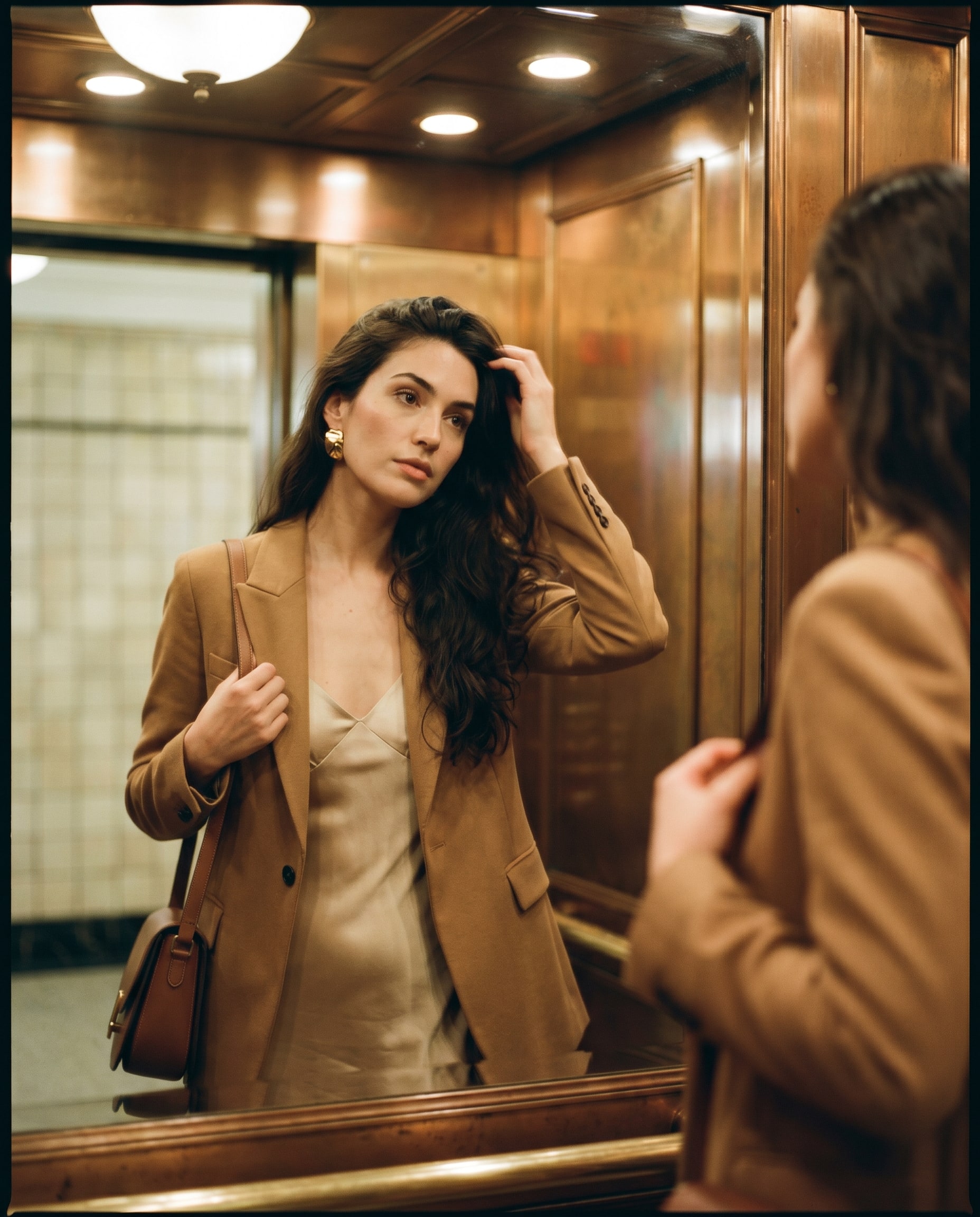 A creator in an elevator checking her reflection, blazer over slip dress, warm overhead light.