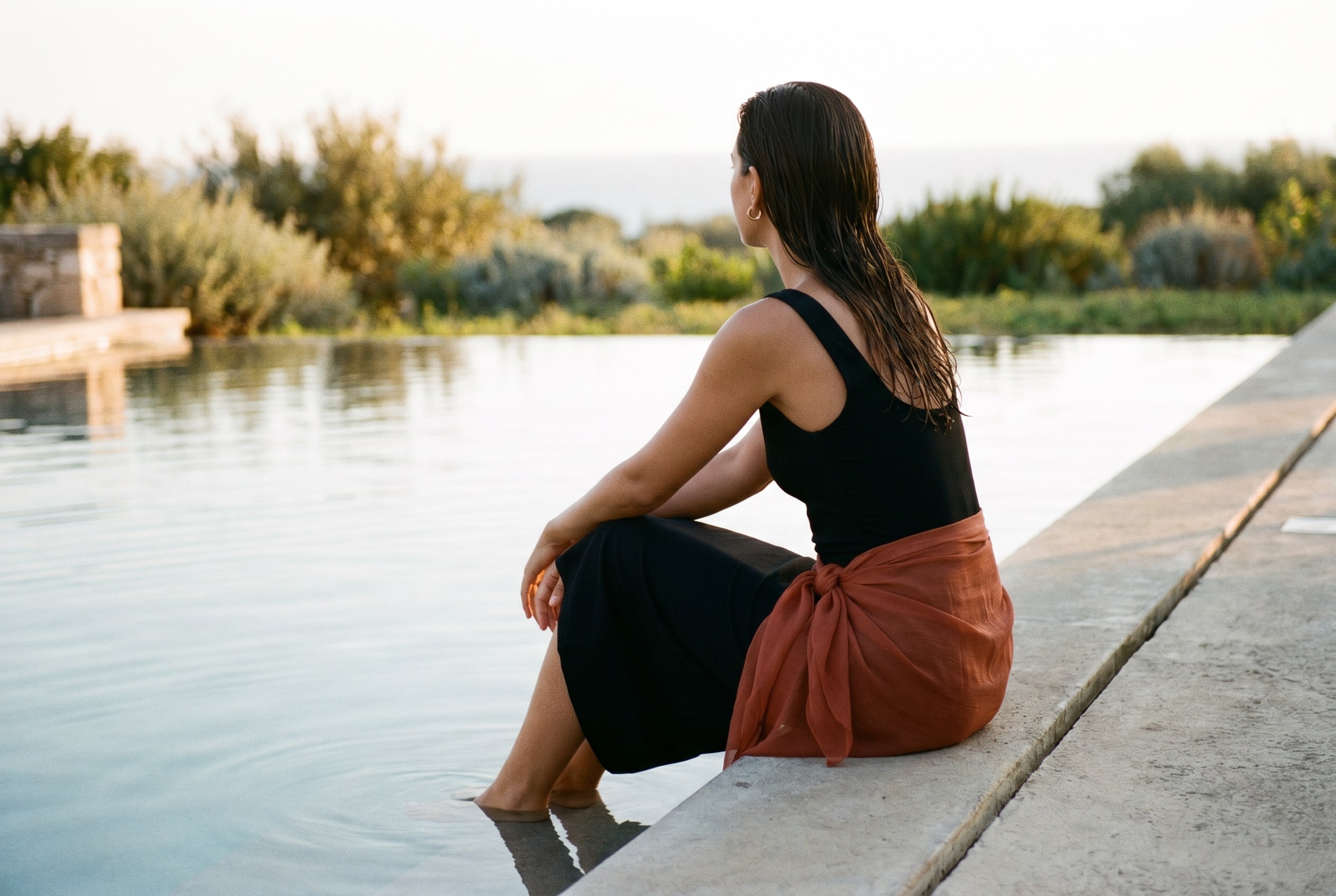 A creator seated at the edge of a pool with feet in the water, one-piece swimsuit, golden hour.