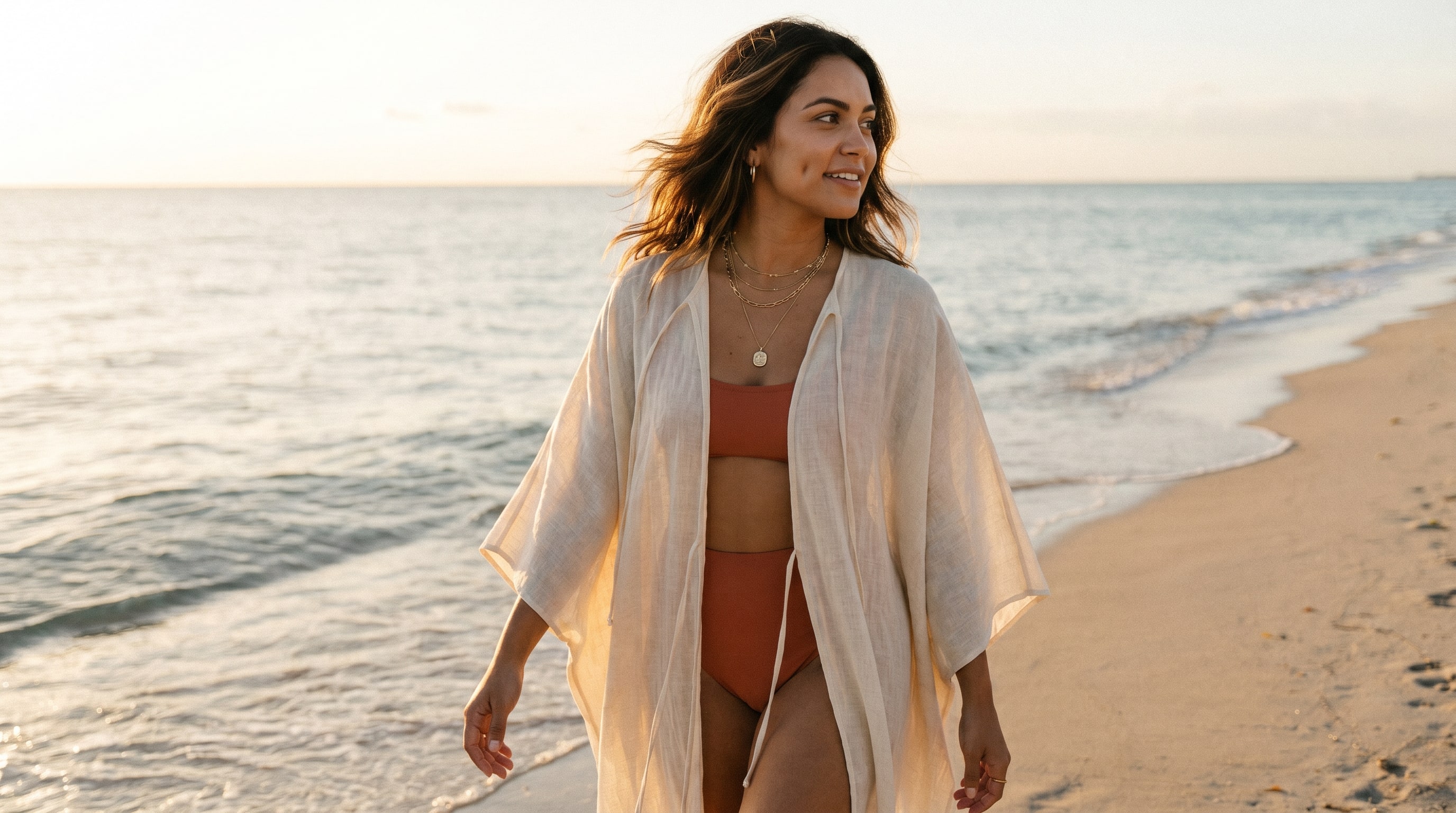 A creator walking along a private beach mid-stride, bikini under a sheer coverup, golden hour.