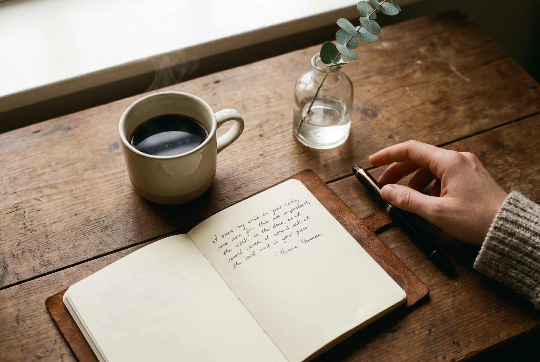 A quiet worktable in early-morning window light — a coffee with steam, a leather notebook open to a handwritten page, a fountain pen, and a small glass vase with a sprig of eucalyptus.